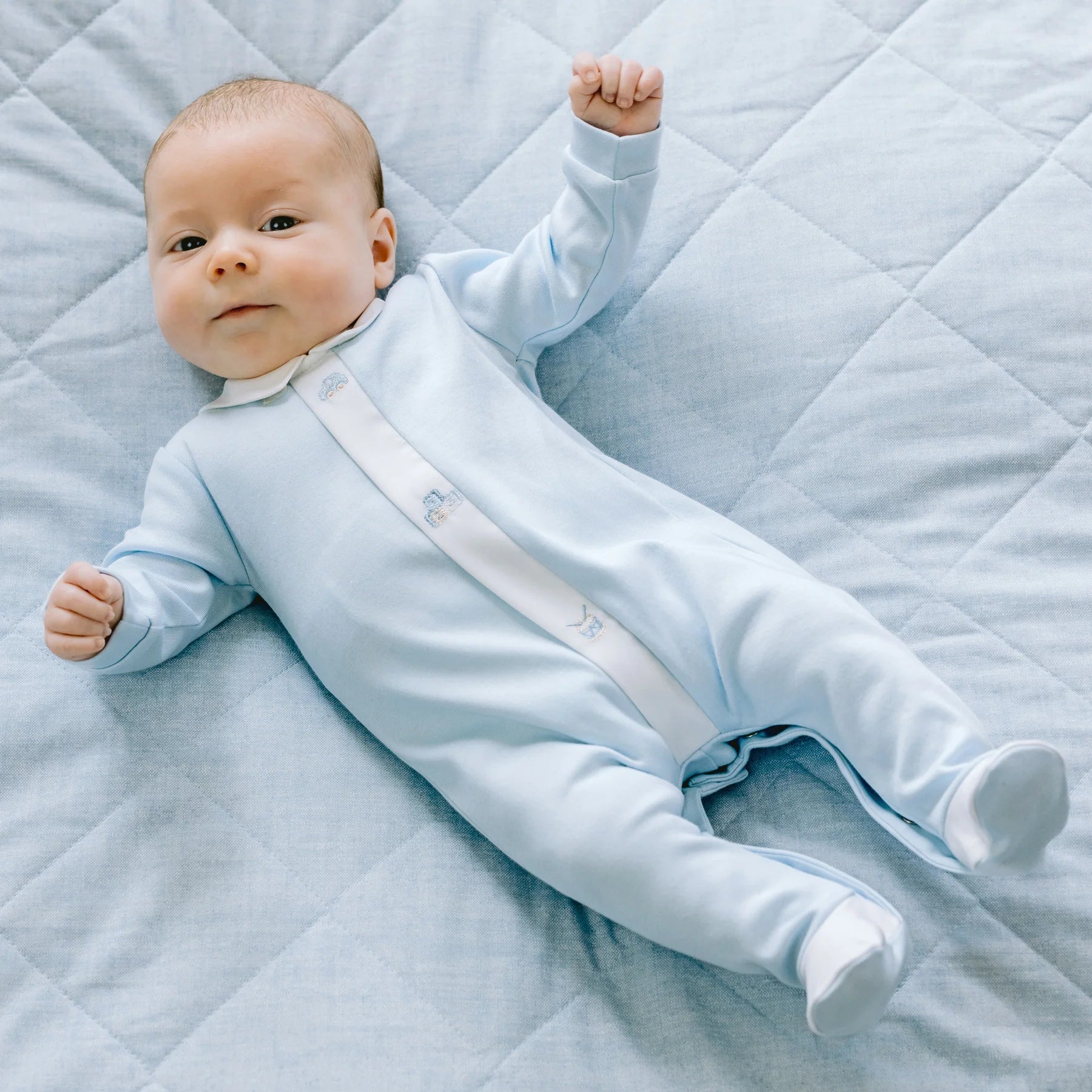 Baby boy lying down in Emile et Rose Kent blue embroidered babygrow.