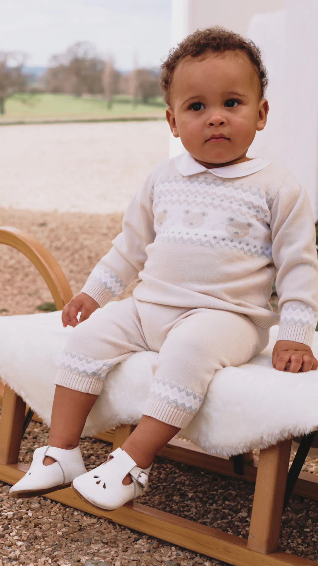 	Baby sitting outdoors in Emile et Rose Kipling knitted teddy outfit set.